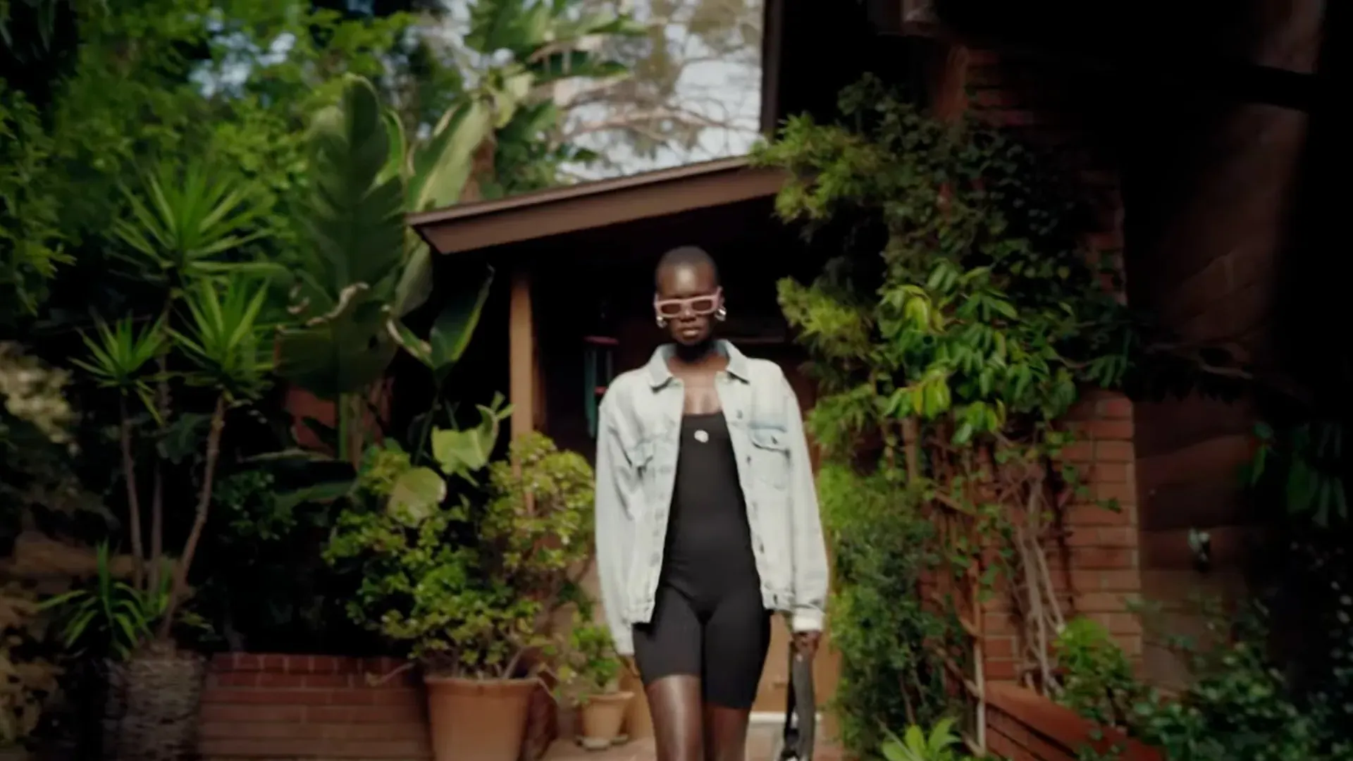 A model in a denim jacket walking out of a house through dense planting, Anine Bing Sport × Carbon38
